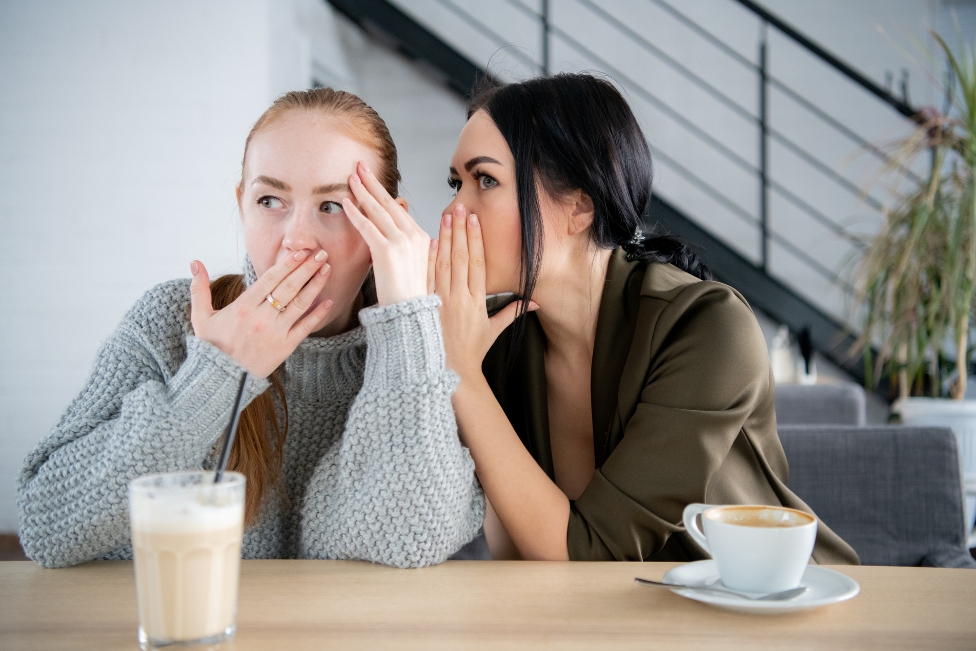 Two women sitting at a table in a café, sharing a secret. The woman on the left has red hair and is wearing a gray sweater, holding her hand over her mouth in surprise. The woman on the right has dark hair, wearing a dark green blouse, whispering into her friend's ear.