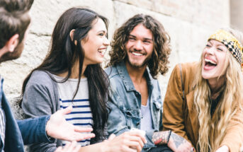 A group of four people, two men and two women, sitting outdoors and laughing together. One woman wears a striped shirt and cardigan, the other has a headband and jacket, one man has long curly hair and tattoos, while the other has short hair and a beard.