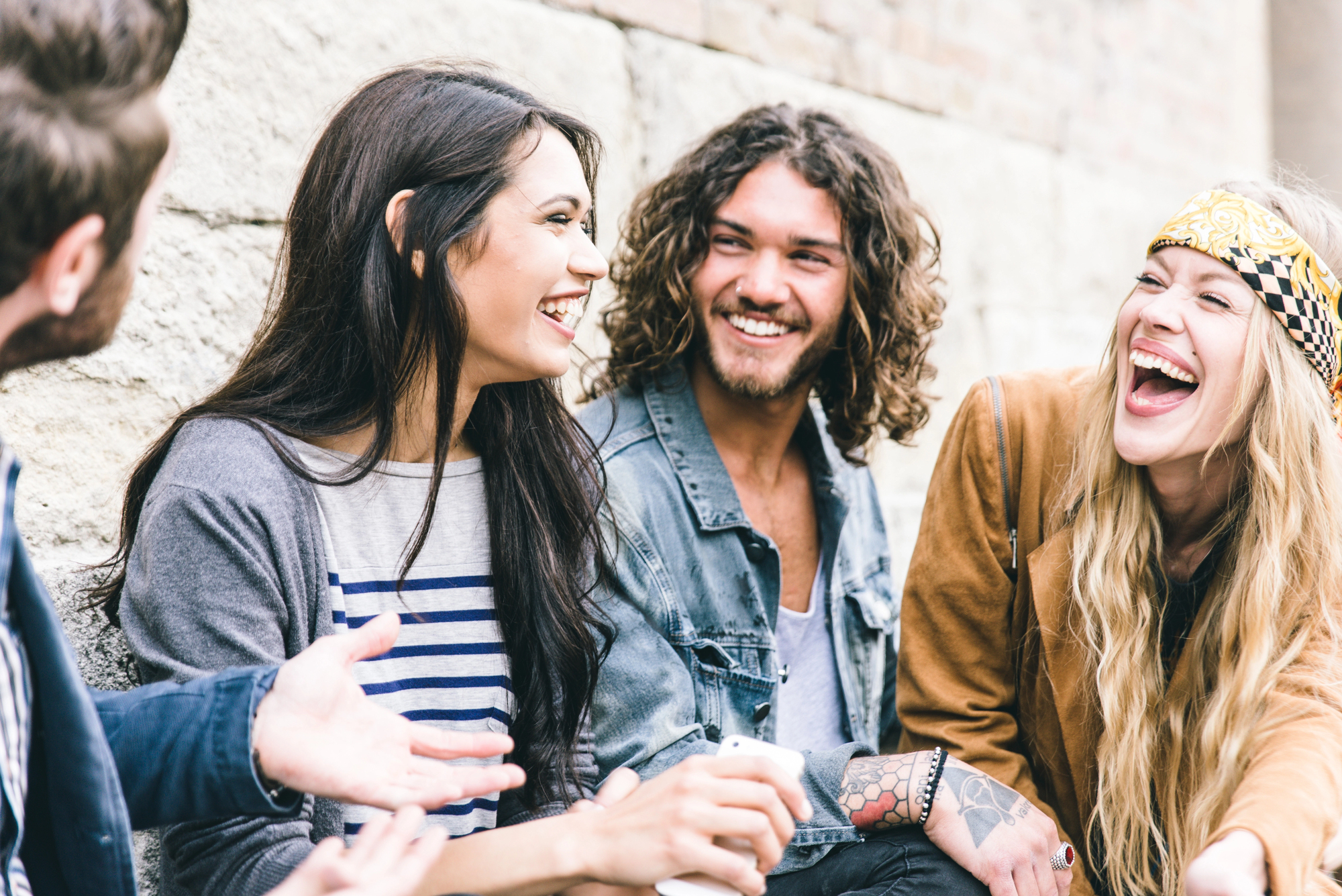 A group of four people, two men and two women, sitting outdoors and laughing together. One woman wears a striped shirt and cardigan, the other has a headband and jacket, one man has long curly hair and tattoos, while the other has short hair and a beard.