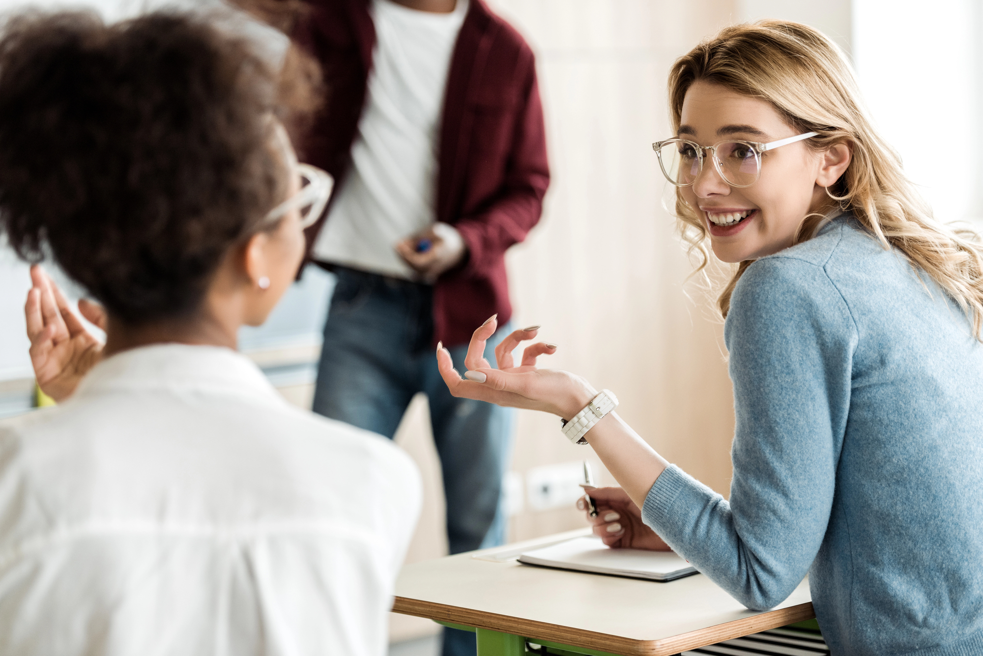 A smiling woman wearing glasses and a blue sweater sits at a desk, engaging in conversation with another woman in a white shirt, who has her back to the camera. A man in a red jacket stands blurred in the background. The scene appears to be in a casual setting.