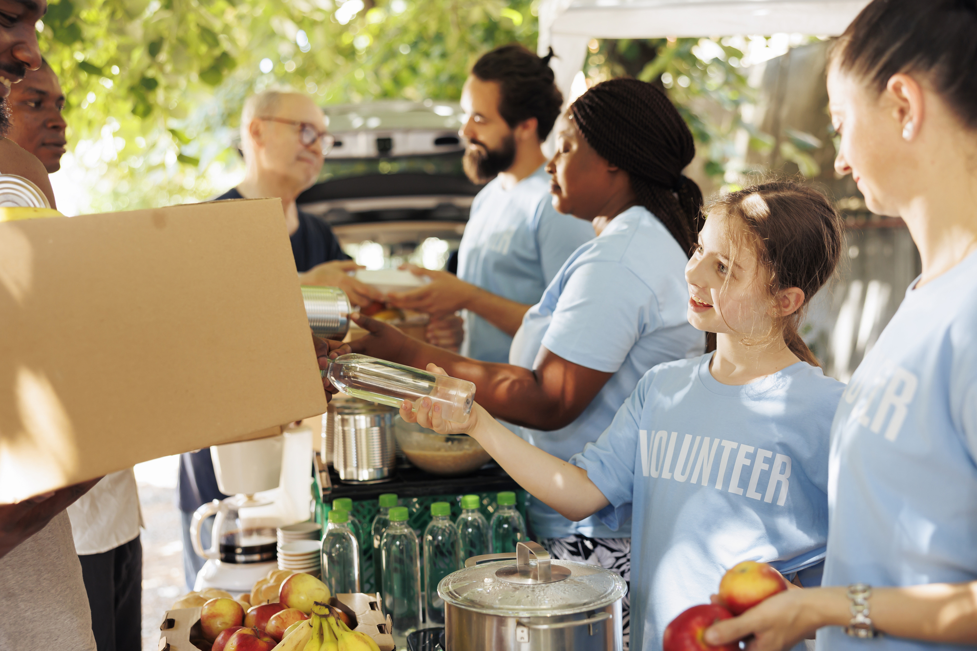 Volunteers distribute food and drinks to people outdoors. A young girl with a "VOLUNTEER" shirt hands a bottle to someone. In the background, other volunteers, also wearing similar shirts, serve the recipients while surrounded by food and supplies.