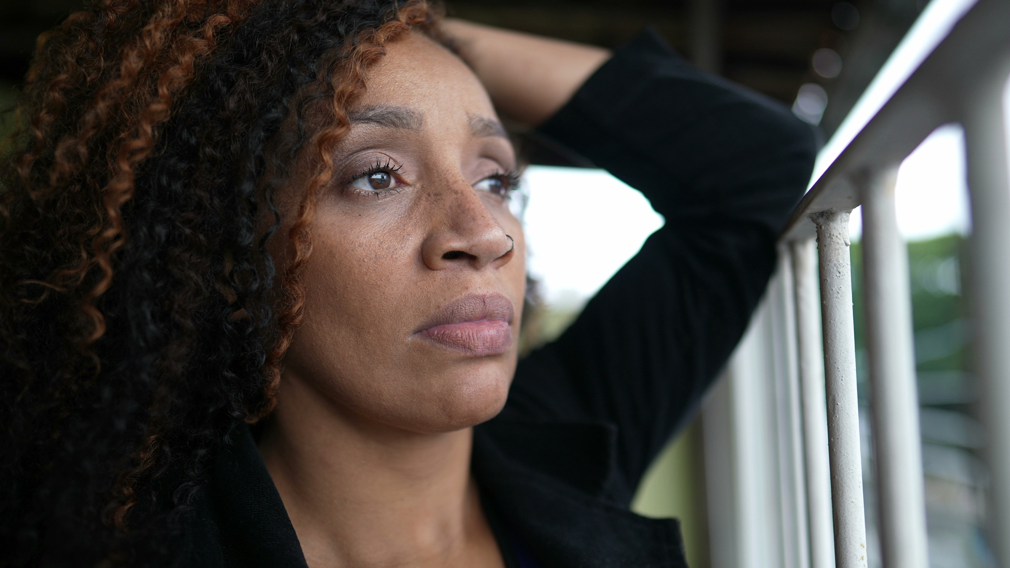 A woman with curly hair gazes thoughtfully out of a window. She has one hand resting behind her head and is wearing a dark jacket. The background is slightly blurred, focusing attention on her contemplative expression.