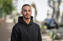 A young man with short hair and a beard, wearing a black hoodie, stands on an outdoor path lined with greenery and parked cars. The background is softly focused, emphasizing the man as the main subject of the image.