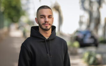 A young man with short hair and a beard, wearing a black hoodie, stands on an outdoor path lined with greenery and parked cars. The background is softly focused, emphasizing the man as the main subject of the image.