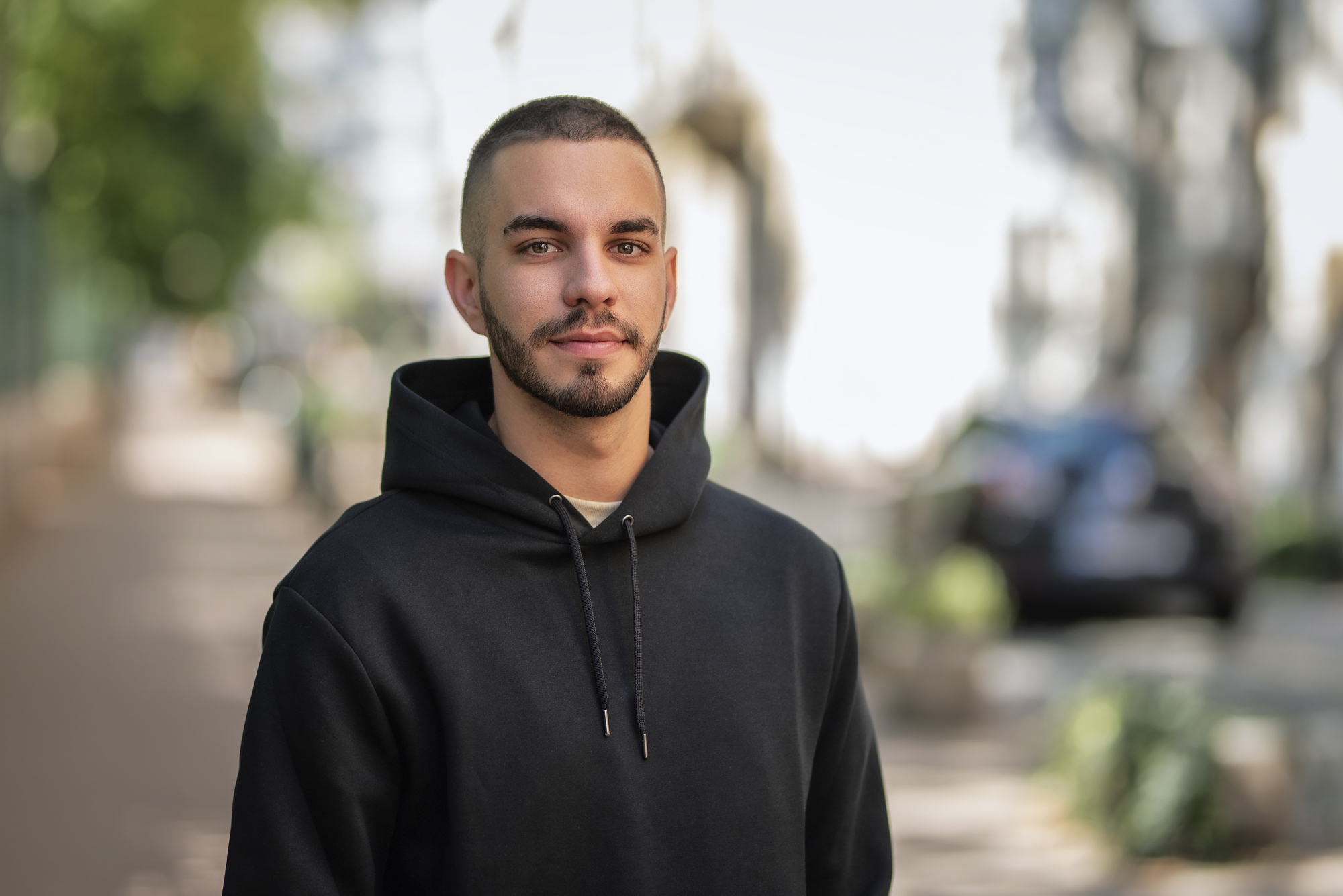 A young man with short hair and a beard, wearing a black hoodie, stands on an outdoor path lined with greenery and parked cars. The background is softly focused, emphasizing the man as the main subject of the image.