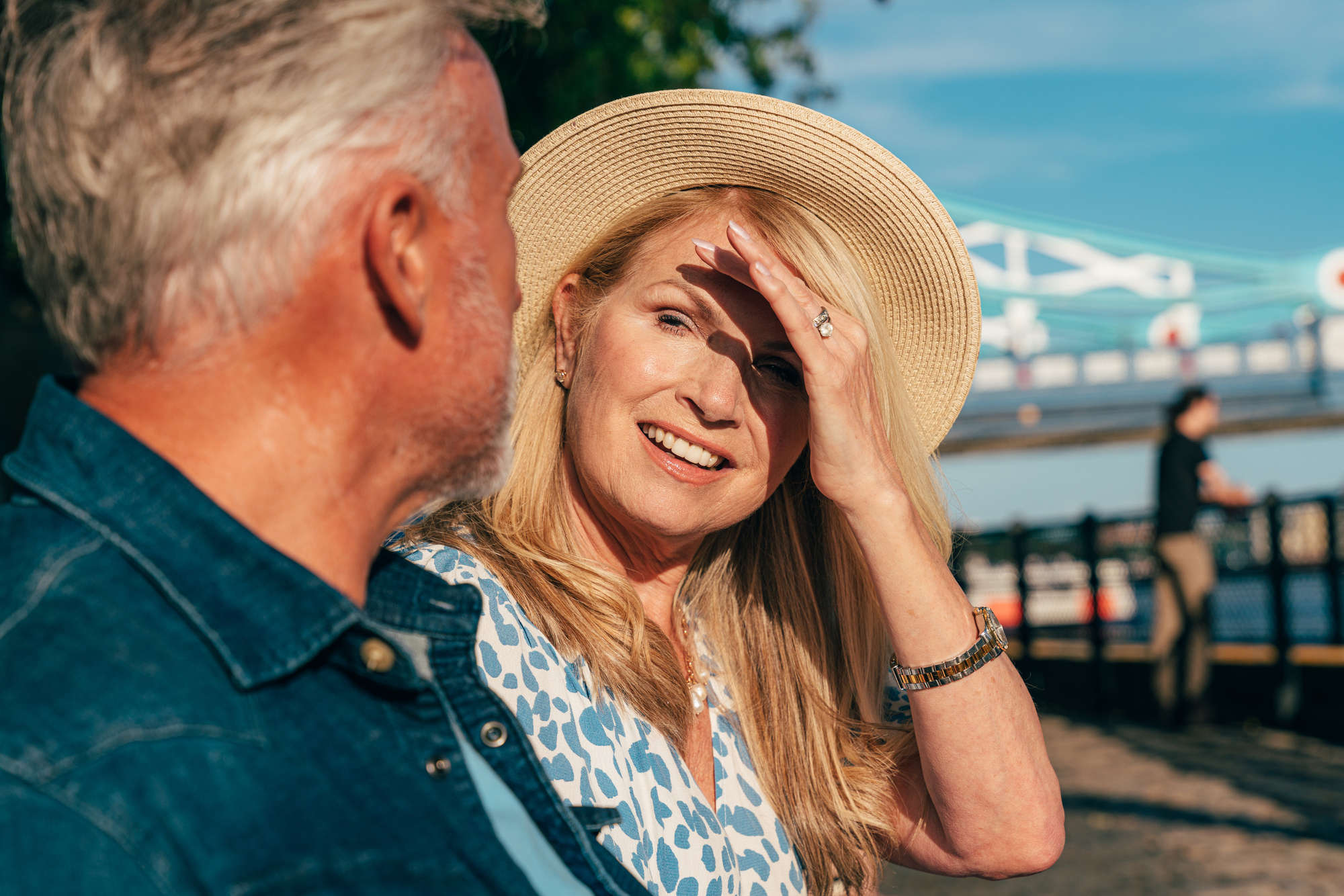 A smiling woman in a sun hat sits outdoors with a man, engaging in conversation. She shields her eyes from the sun with one hand. Behind them is a bridge with blue and white structures. The atmosphere is bright and serene.