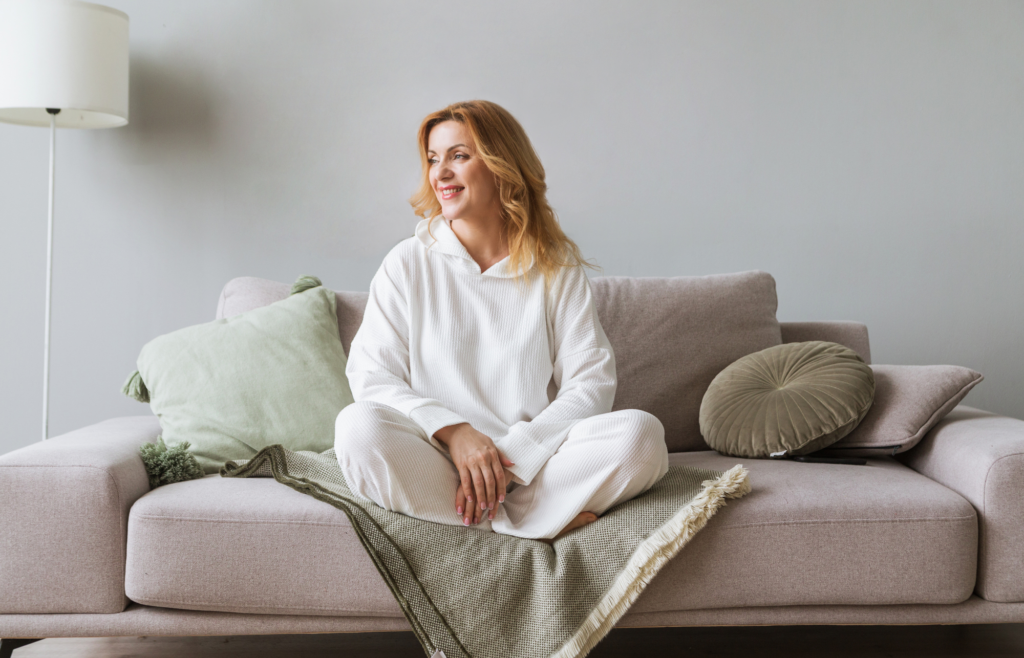 A woman with long blonde hair, wearing a white hoodie and light-colored pants, sits cross-legged on a beige sofa. She looks to the side with a smile. The sofa is adorned with green and beige pillows and a green throw blanket. A white lamp stands in the background.