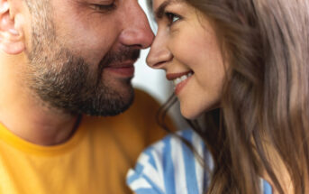 A close-up of a smiling couple facing each other. The man has a beard and is wearing a yellow shirt, while the woman has long brown hair and is wearing a blue and white striped shirt. They appear happy and content, with their noses nearly touching.