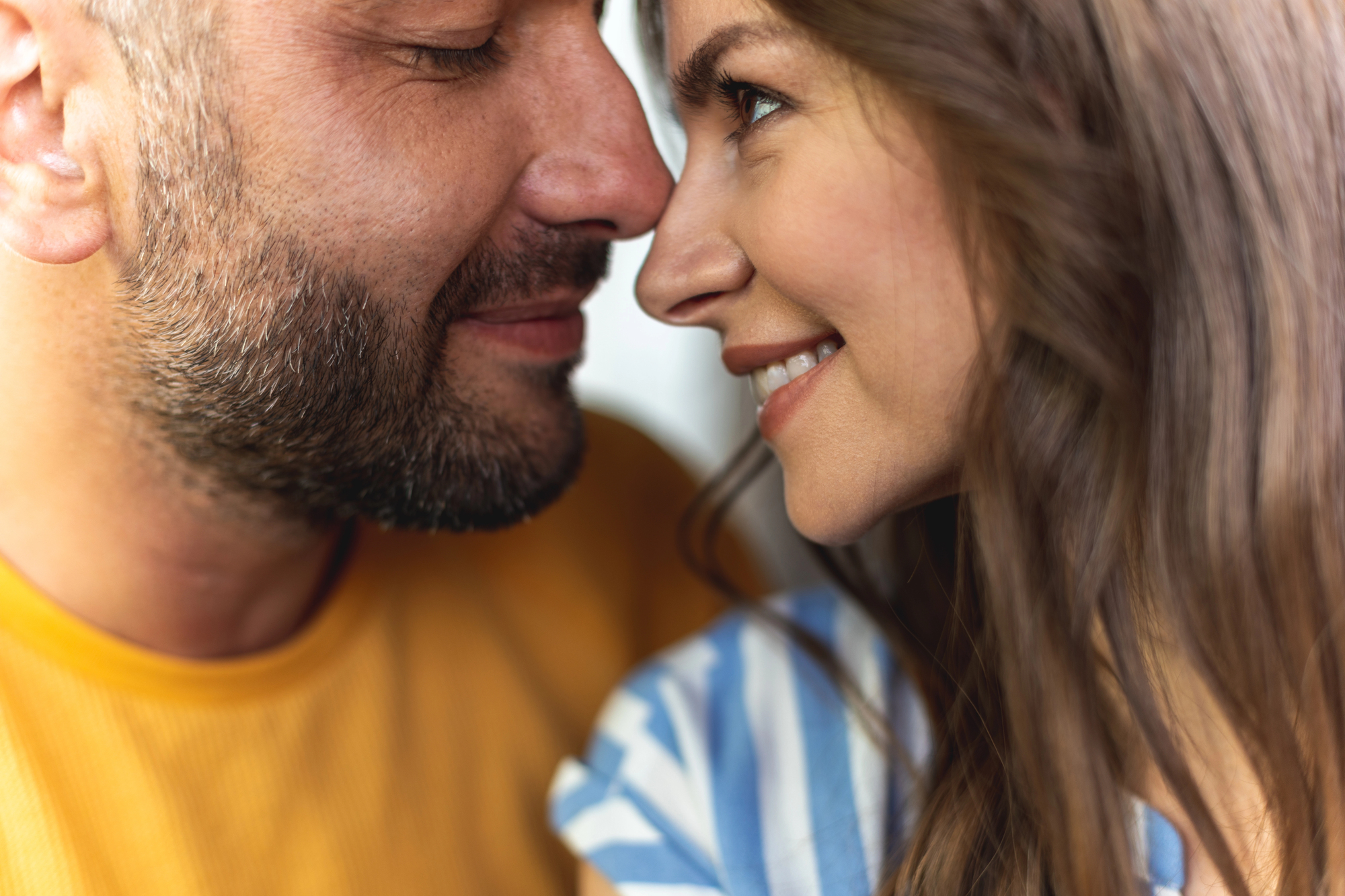 A close-up of a smiling couple facing each other. The man has a beard and is wearing a yellow shirt, while the woman has long brown hair and is wearing a blue and white striped shirt. They appear happy and content, with their noses nearly touching.