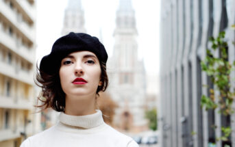 A person with red lipstick and a black beret stands outdoors in front of a blurred background featuring tall buildings, including one with spires. They are wearing a white turtleneck, and their expression is neutral.