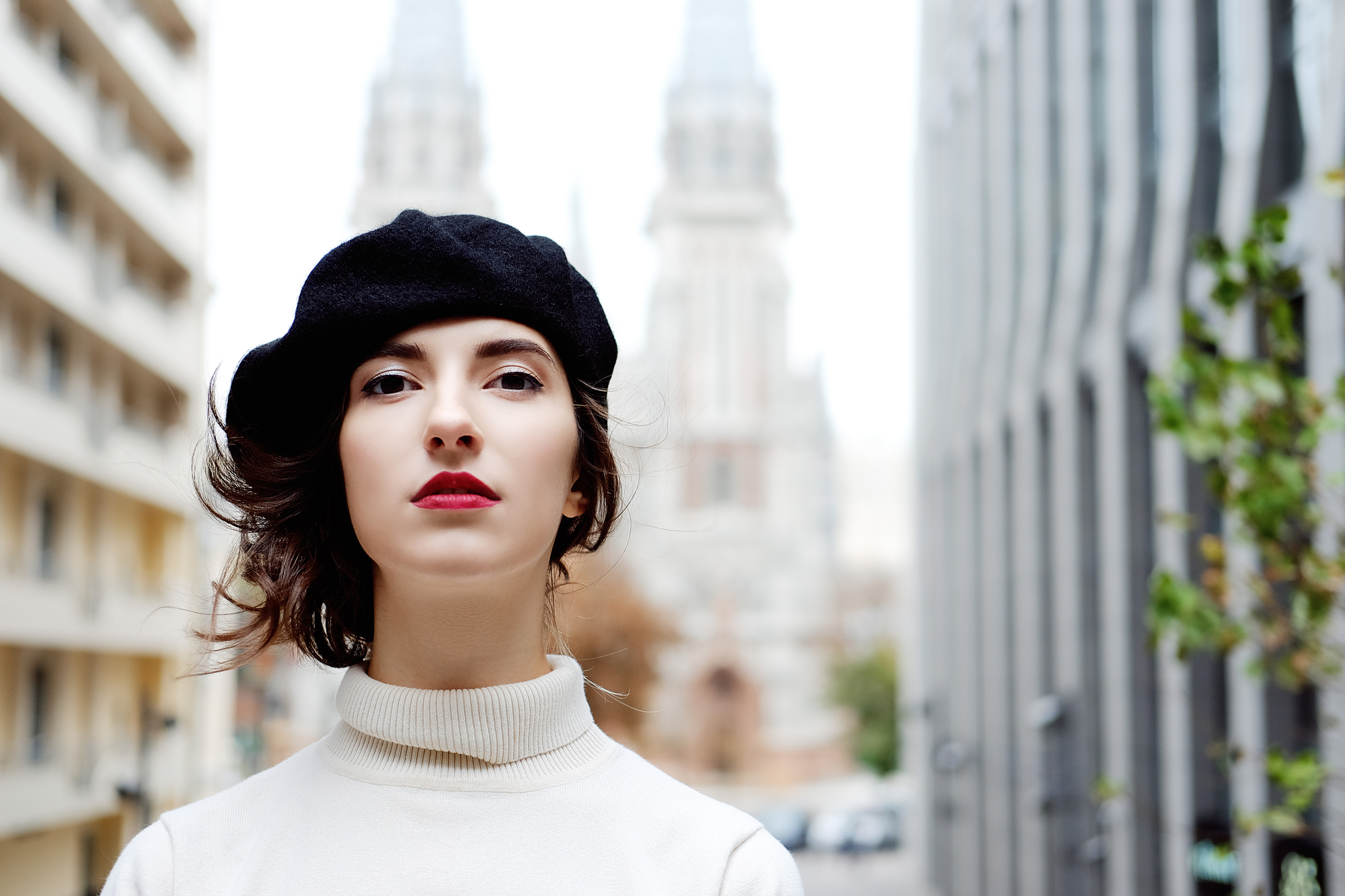 A person with red lipstick and a black beret stands outdoors in front of a blurred background featuring tall buildings, including one with spires. They are wearing a white turtleneck, and their expression is neutral.