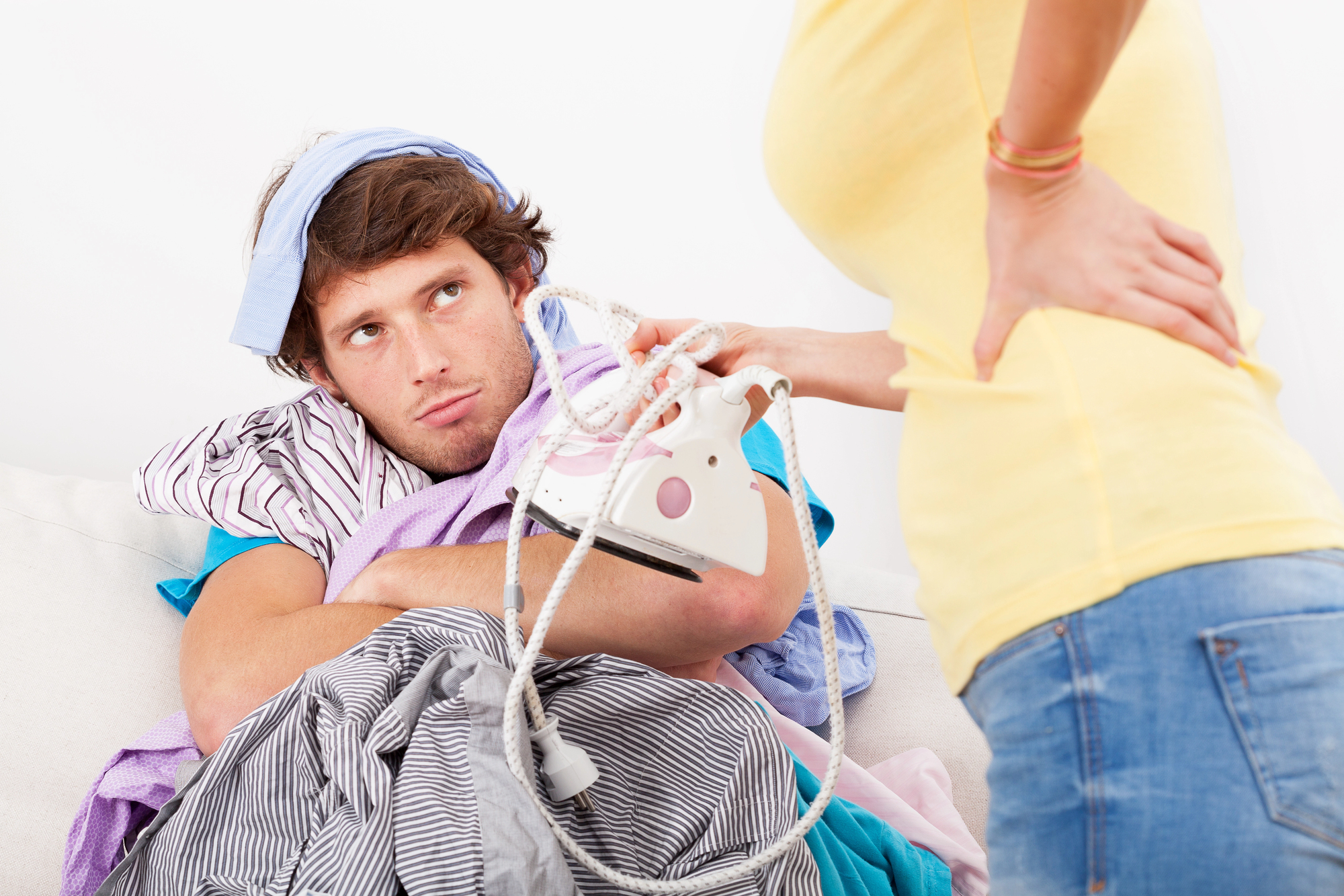 A man sits on a couch with a frustrated expression, holding an iron and surrounded by a disorganized pile of clothes. A woman stands in front of him with her hands on her hips, only her torso visible, wearing a yellow top and jeans.