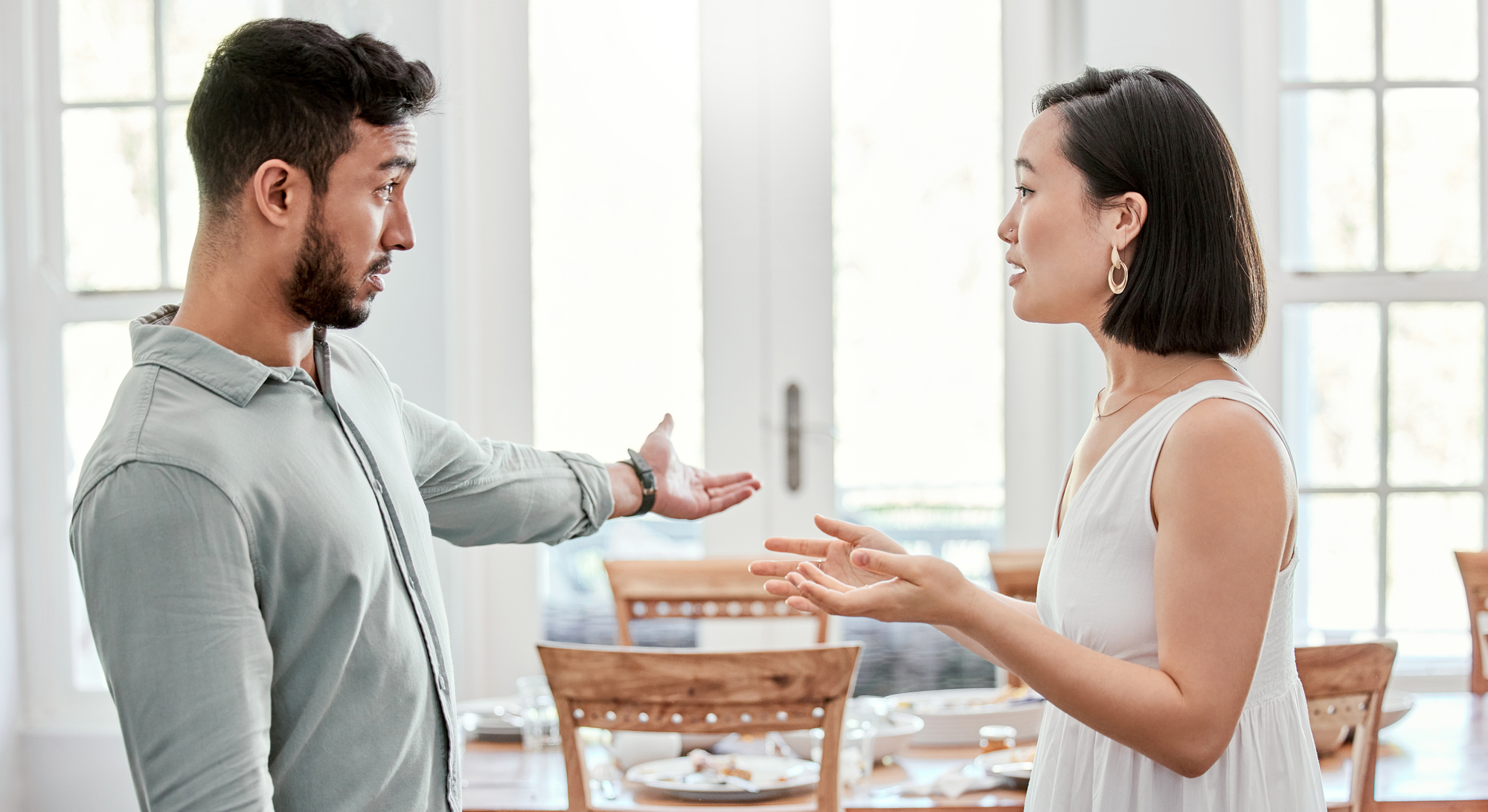 A man and a woman are standing in a bright room, engaged in a heated conversation. The man has an expressive gesture with one hand extended, while the woman is gesturing with both hands. They appear to be in a dining area with a table set in the background.