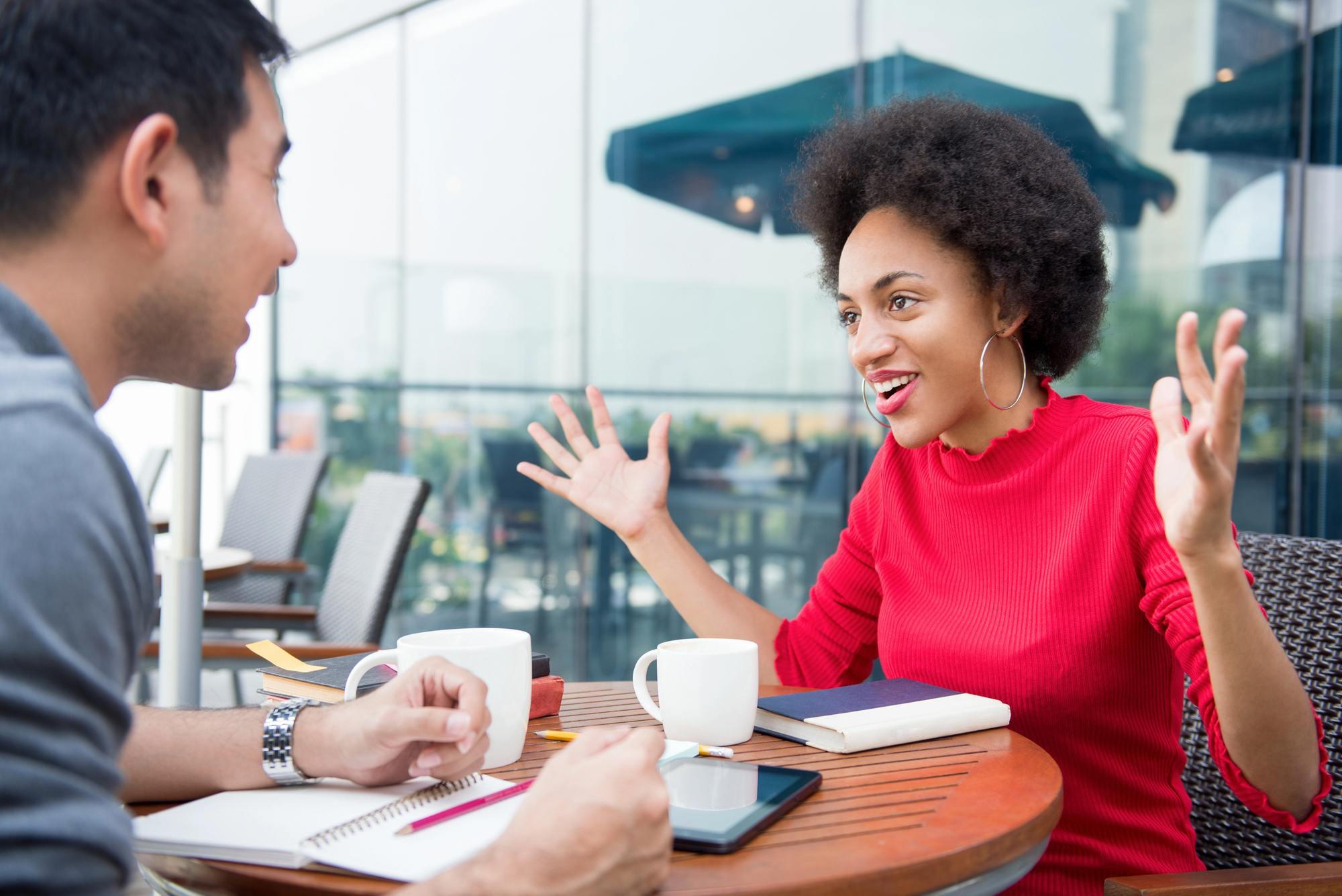 Two people are seated at an outdoor café table engaged in an animated conversation. One person is wearing a red sweater and gesturing with hands raised. The table has notebooks, a tablet, and coffee cups. The background shows a glass building and other café furniture.