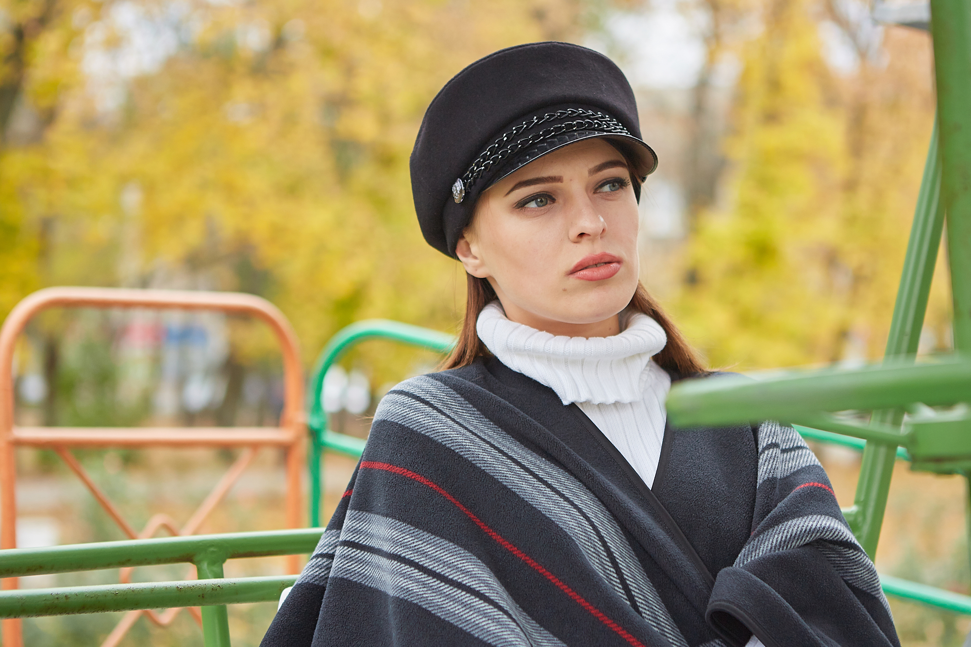 A woman with a neutral expression is sitting outdoors. She is wearing a black hat with chain detail, a white turtleneck sweater, and a dark shawl with red and grey stripes. The background features blurred autumn trees and green and orange metal bars.