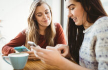 Two women are sitting at a table in a cozy café, both focused on their smartphones. One is showing something on her phone to the other, who is smiling. A cup of coffee is on the table in front of them, creating a casual, relaxed atmosphere.