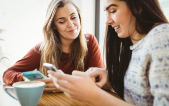 Two women are sitting at a table in a cozy café, both focused on their smartphones. One is showing something on her phone to the other, who is smiling. A cup of coffee is on the table in front of them, creating a casual, relaxed atmosphere.