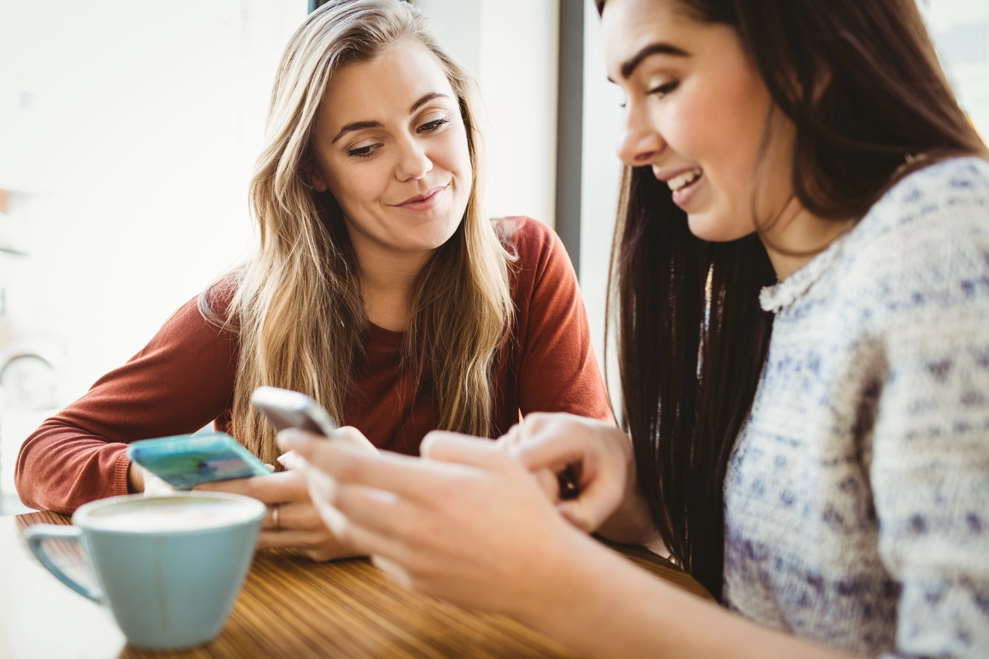 Two women are sitting at a table in a cozy café, both focused on their smartphones. One is showing something on her phone to the other, who is smiling. A cup of coffee is on the table in front of them, creating a casual, relaxed atmosphere.