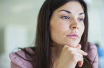 A woman with long dark hair and a thoughtful expression rests her chin on her hand while gazing into the distance. She is wearing a light pink top and appears to be in a well-lit indoor setting.