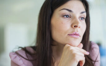 A woman with long dark hair and a thoughtful expression rests her chin on her hand while gazing into the distance. She is wearing a light pink top and appears to be in a well-lit indoor setting.