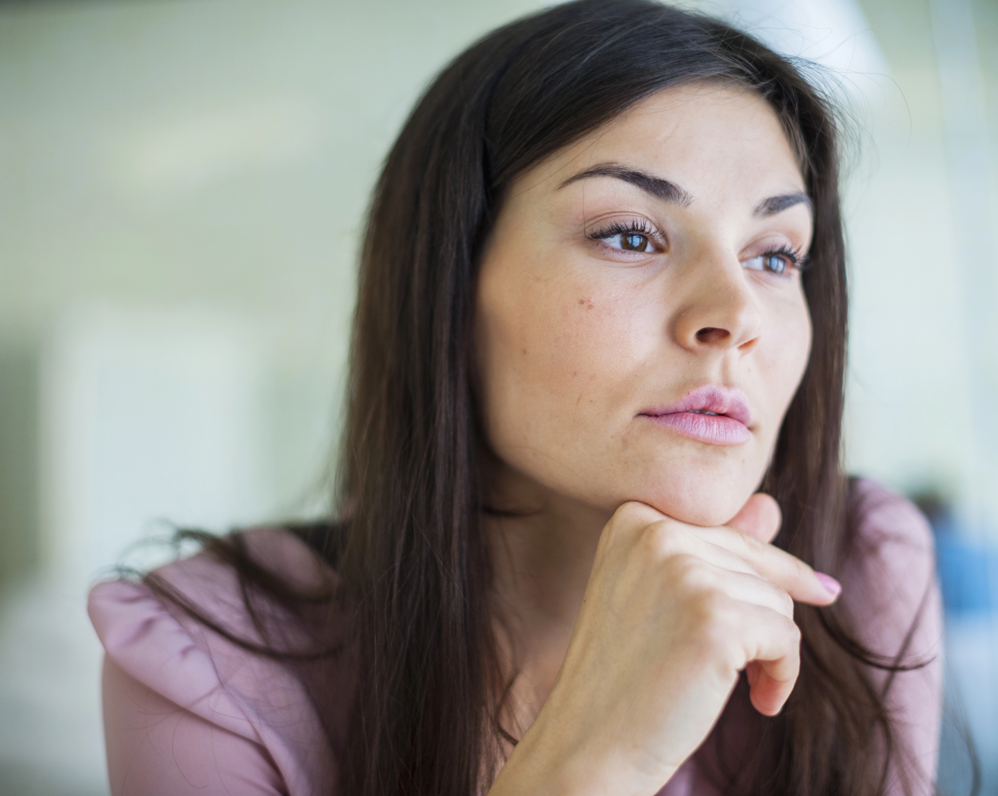 A woman with long dark hair and a thoughtful expression rests her chin on her hand while gazing into the distance. She is wearing a light pink top and appears to be in a well-lit indoor setting.