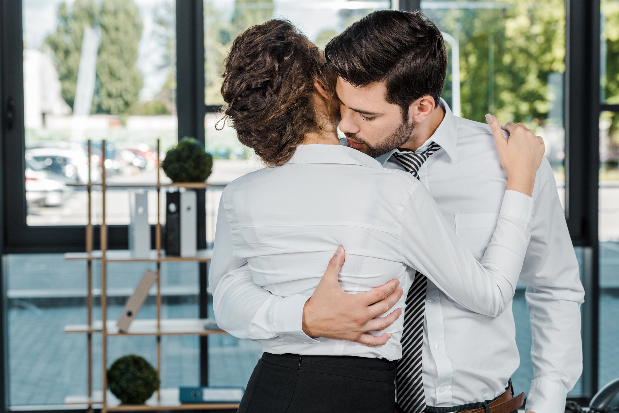 A man and woman in business attire embrace and kiss in an office setting. The man places one hand on the woman's back while the other holds her waist. The woman has her arm around his shoulder. A shelf and large windows with a view of trees and parked cars are behind them.