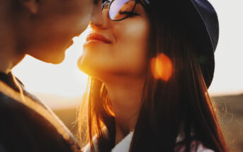 A close-up of a couple's affectionate moment. The woman, wearing glasses and a hat, smiles gently while leaning towards the man, whose face is partially visible. Sunlight creates a warm, golden glow with lens flare effects in the background.