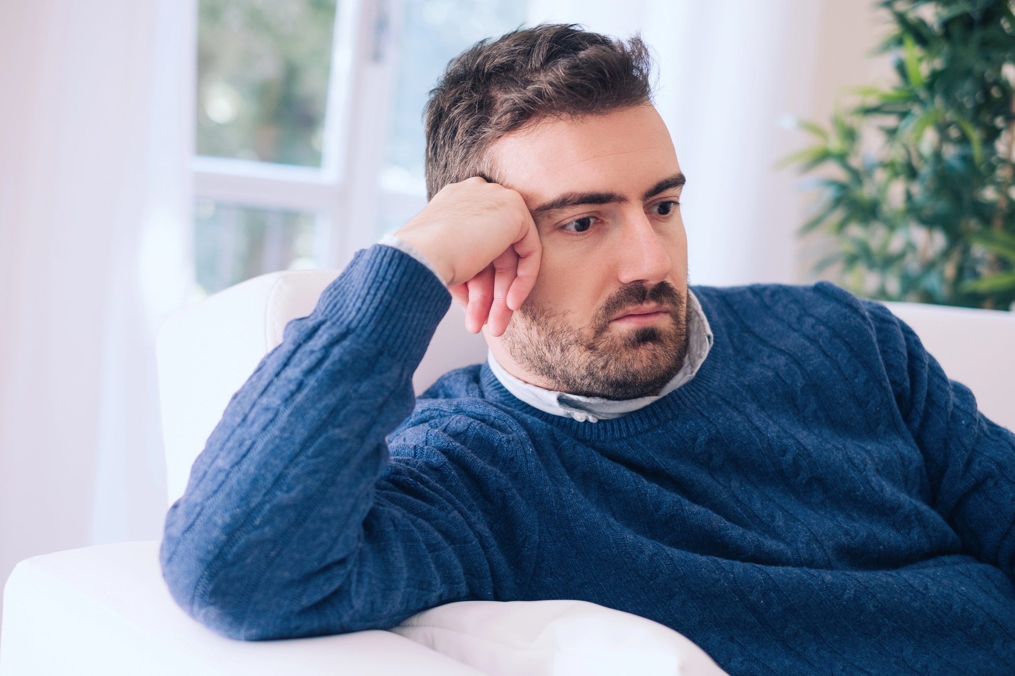 A man with short dark hair and a beard, wearing a blue sweater, rests his head on his hand while sitting on a white sofa. He appears deep in thought, with a window and a green plant visible in the background.