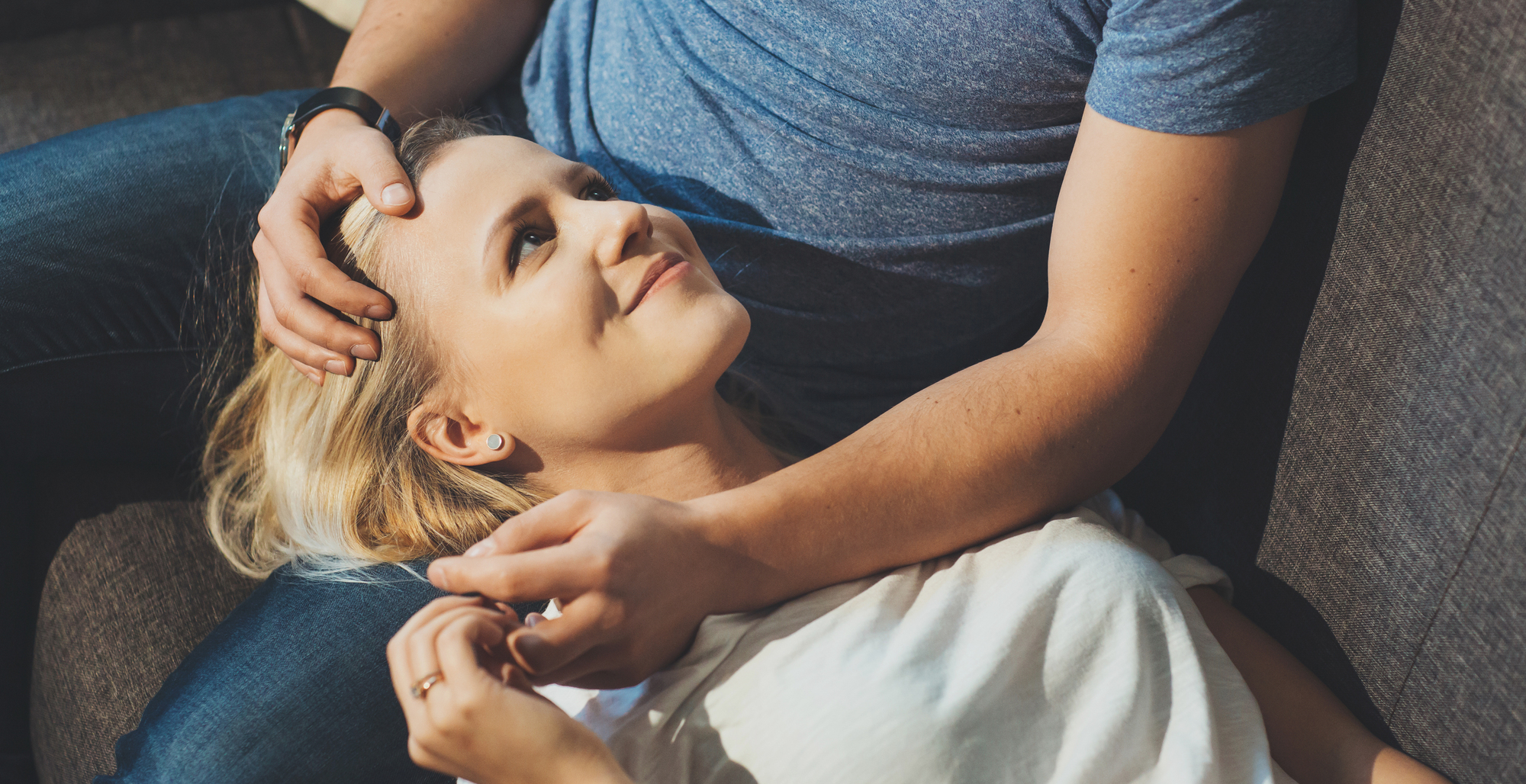 A person with long, blonde hair smiles while lying on a couch with their head resting on another person's lap. The person whose lap is being used is wearing a blue shirt and jeans, gently holding the head of the person lying down.