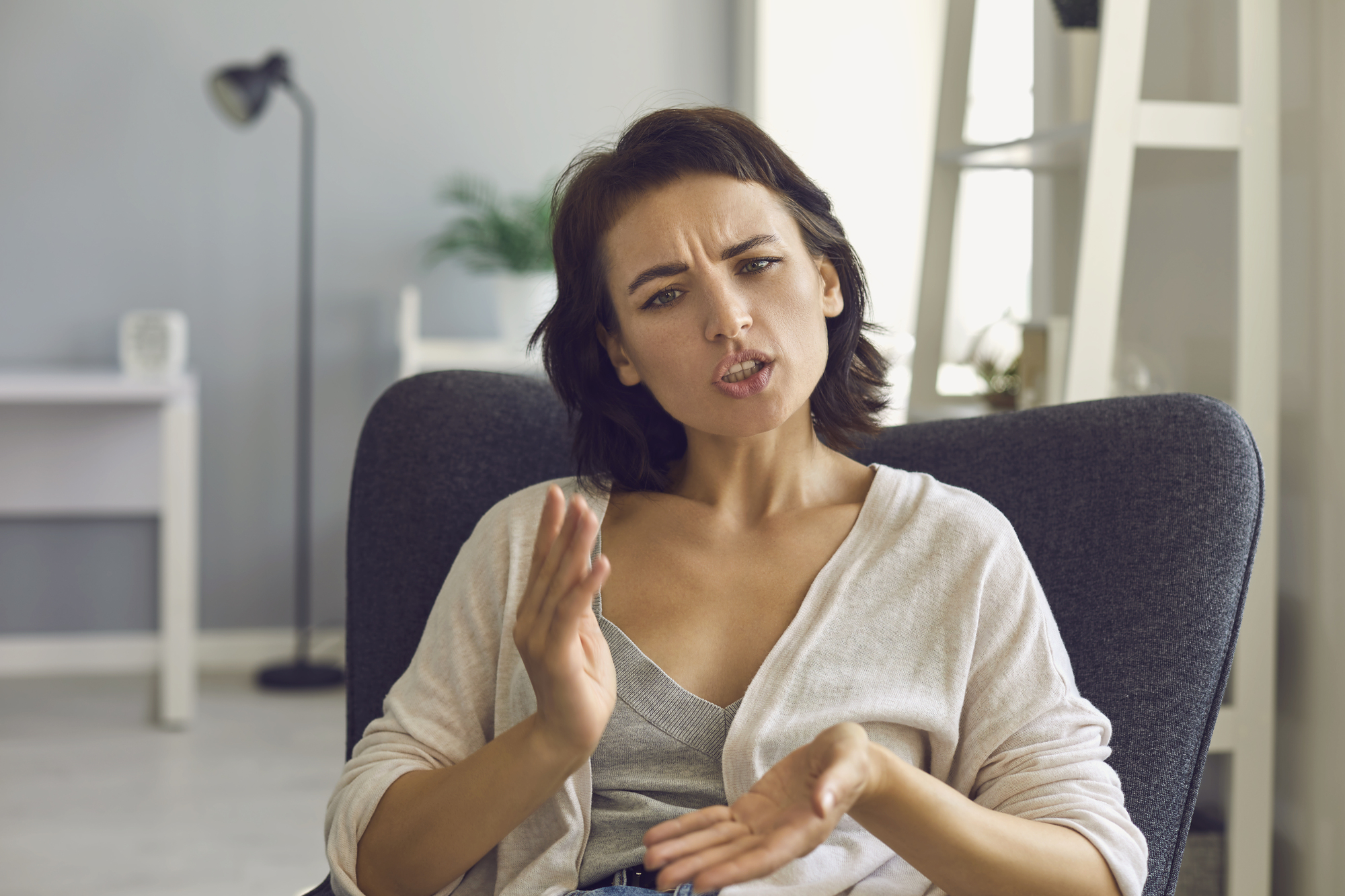 A woman with short brown hair is sitting in a chair, gesturing with her hands while looking intently at someone. She appears to be engaged in a serious conversation. The background shows a blurred interior with soft lighting, a lamp, and a piece of furniture.