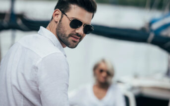 A man with dark hair and a beard, wearing sunglasses and a white shirt, looks over his shoulder. A woman, slightly out of focus in the background, also wearing a white shirt, is seated. They are outdoors, possibly on a boat or near water.