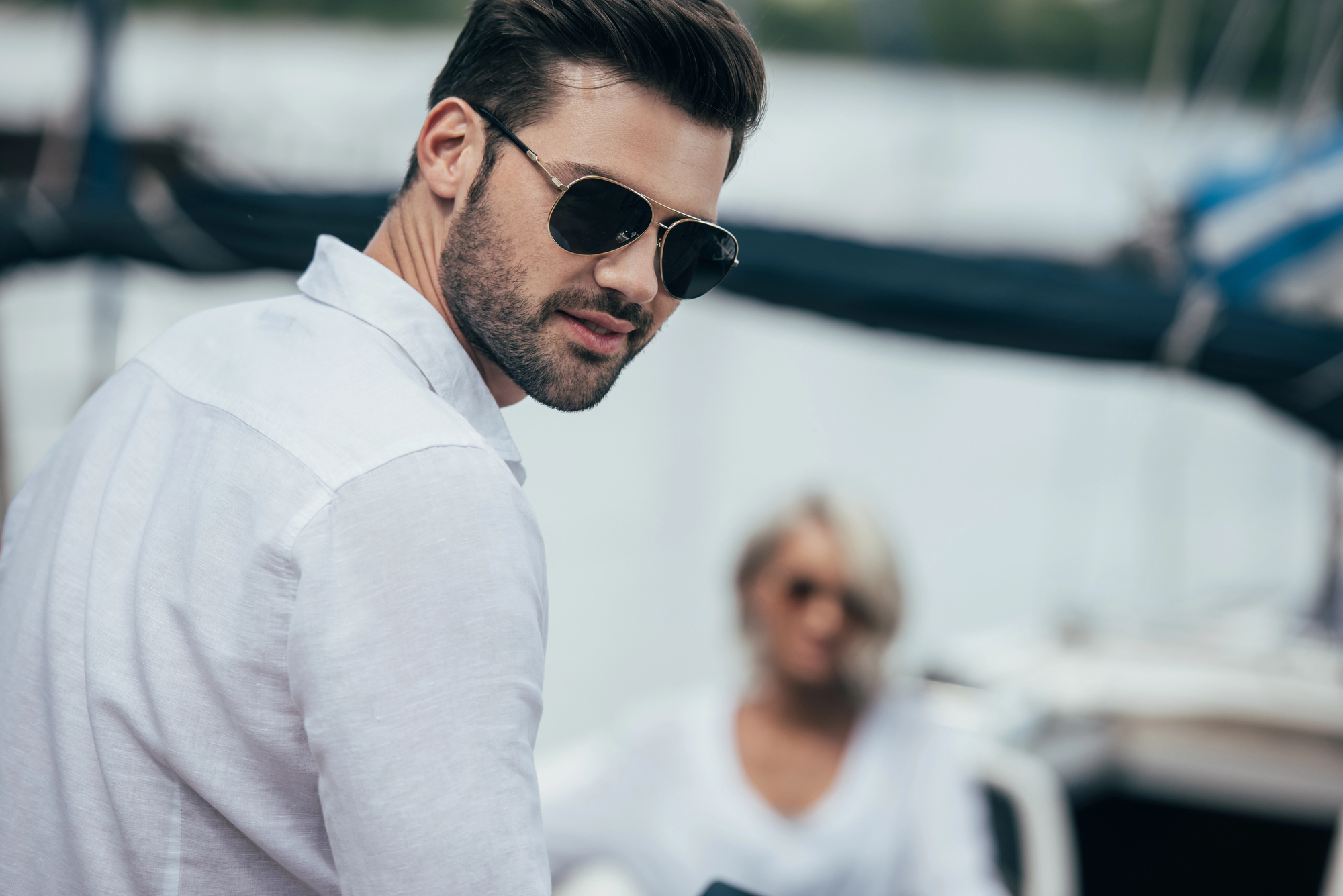 A man with dark hair and a beard, wearing sunglasses and a white shirt, looks over his shoulder. A woman, slightly out of focus in the background, also wearing a white shirt, is seated. They are outdoors, possibly on a boat or near water.