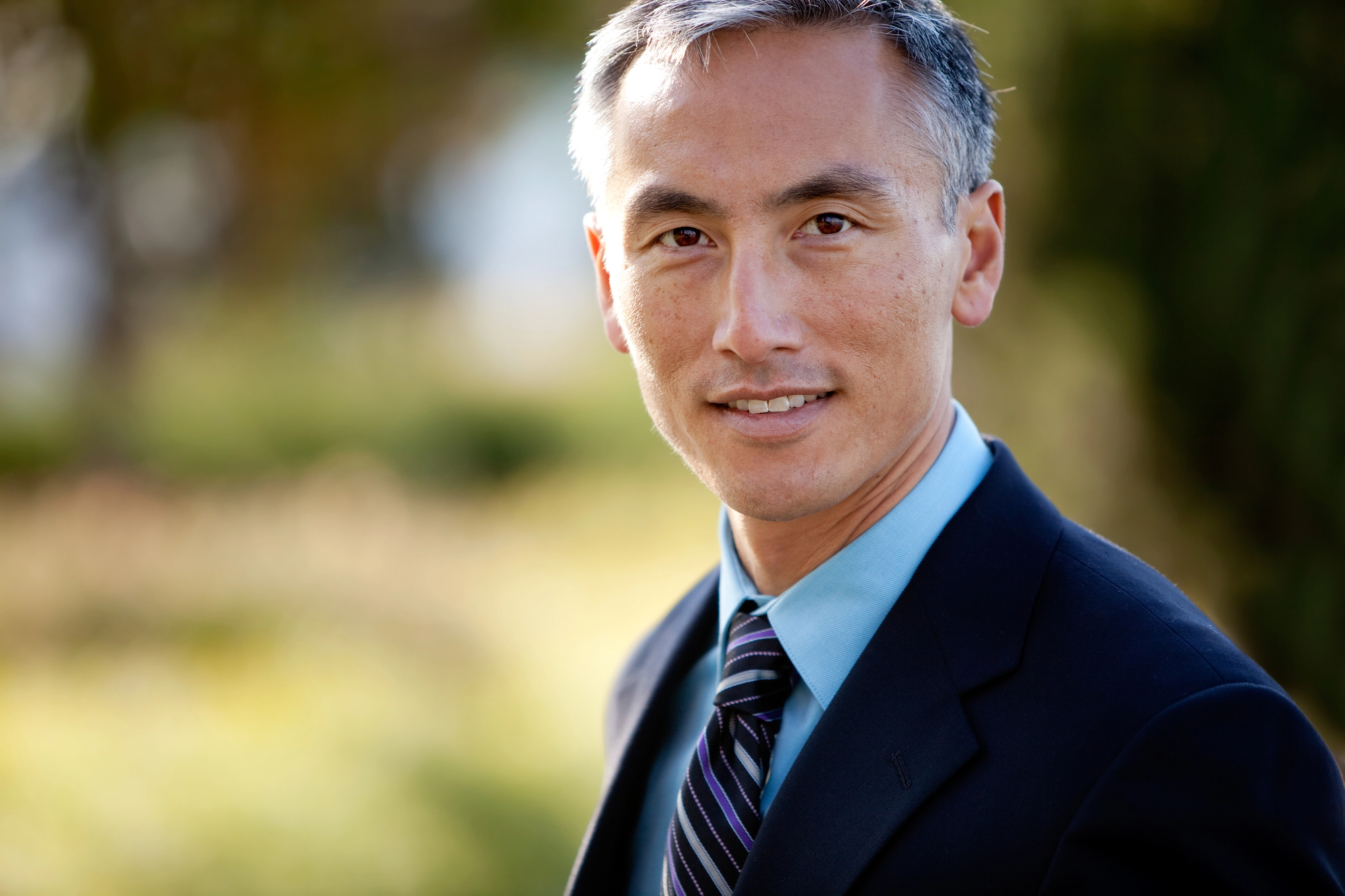 A man with short gray hair, wearing a black suit, blue shirt, and striped tie. He is standing outdoors with a blurred natural background, looking directly at the camera with a slight smile.