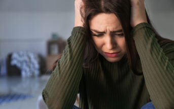 A young woman with long brown hair, wearing a green ribbed sweater, sits indoors with her hands on her head. She appears distressed, furrowing her brows and looking down. The background is blurred, showing furniture and a blanket on the floor.