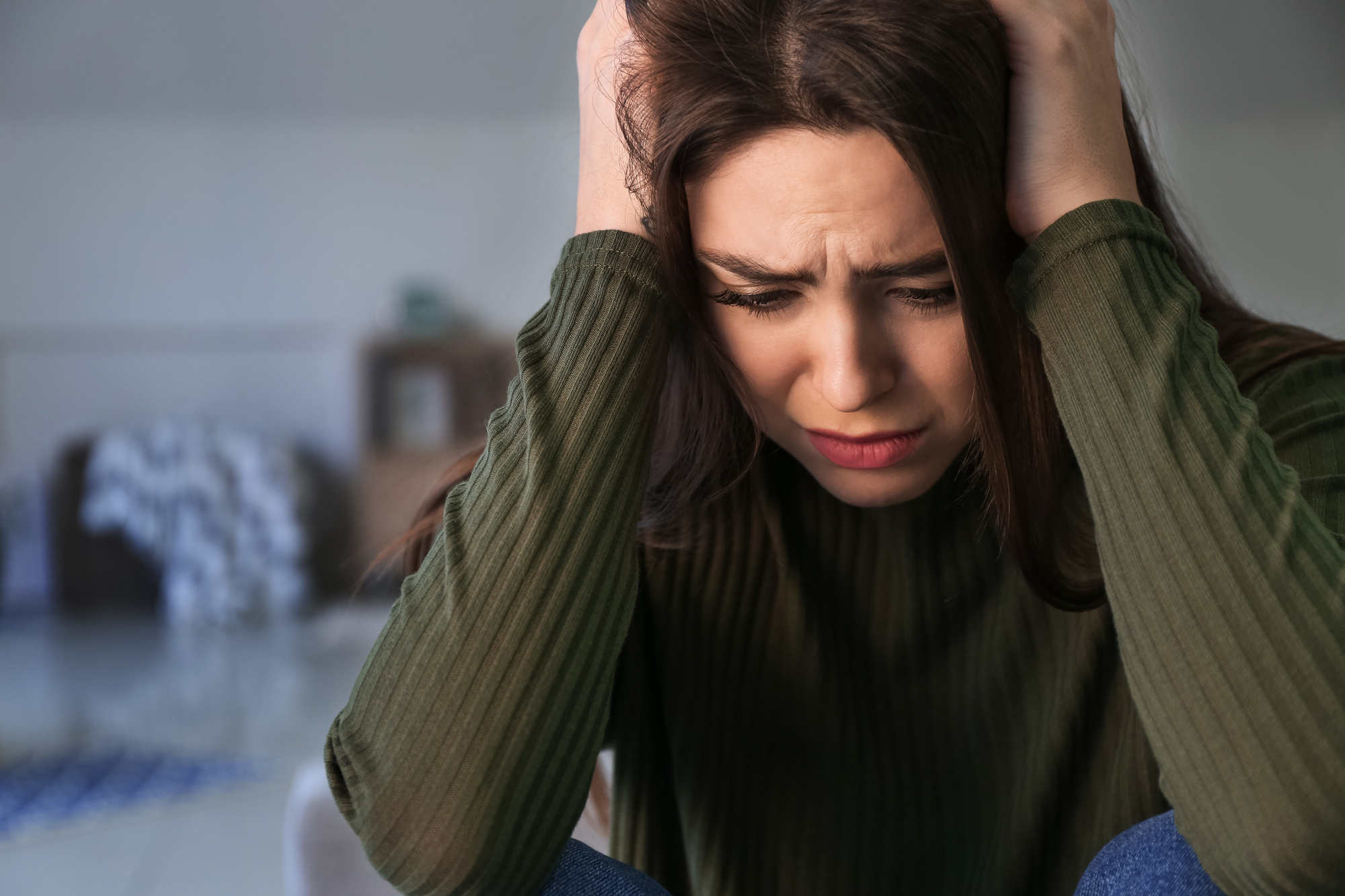 A young woman with long brown hair, wearing a green ribbed sweater, sits indoors with her hands on her head. She appears distressed, furrowing her brows and looking down. The background is blurred, showing furniture and a blanket on the floor.