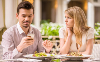 A man and a woman are sitting at a table in a restaurant. The man is looking at his smartphone while the woman, with blonde hair, is resting her chin on her hand, gazing at him. Both have plates of food in front of them on the table.