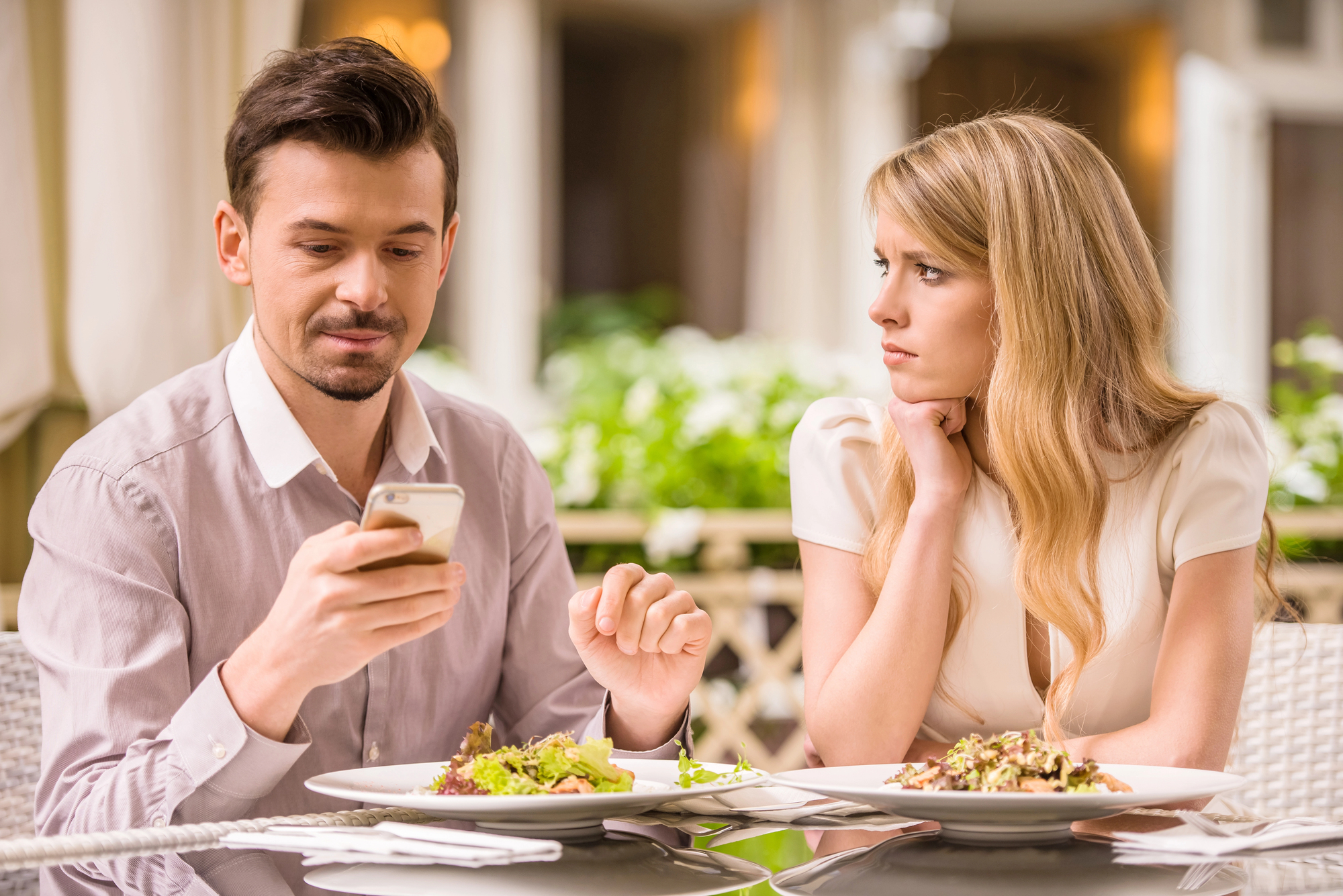 A man and a woman are sitting at a table in a restaurant. The man is looking at his smartphone while the woman, with blonde hair, is resting her chin on her hand, gazing at him. Both have plates of food in front of them on the table.