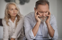 An older man with a gray beard sits with his head in his hands, looking distressed. An older woman with long blonde hair, wearing a white blouse, sits beside him with a concerned expression. The background is neutral and out of focus.