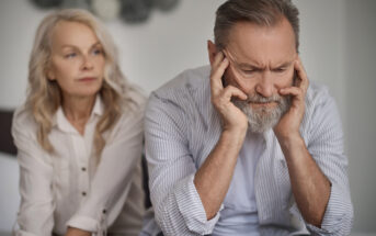 An older man with a gray beard sits with his head in his hands, looking distressed. An older woman with long blonde hair, wearing a white blouse, sits beside him with a concerned expression. The background is neutral and out of focus.