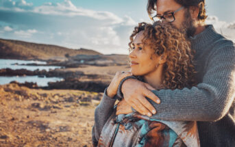 A couple stands close together on a rocky beach with clear blue skies and water in the background. The man, in glasses and a gray sweater, hugs the woman from behind. The woman, with curly hair, smiles contently, both looking towards the horizon.