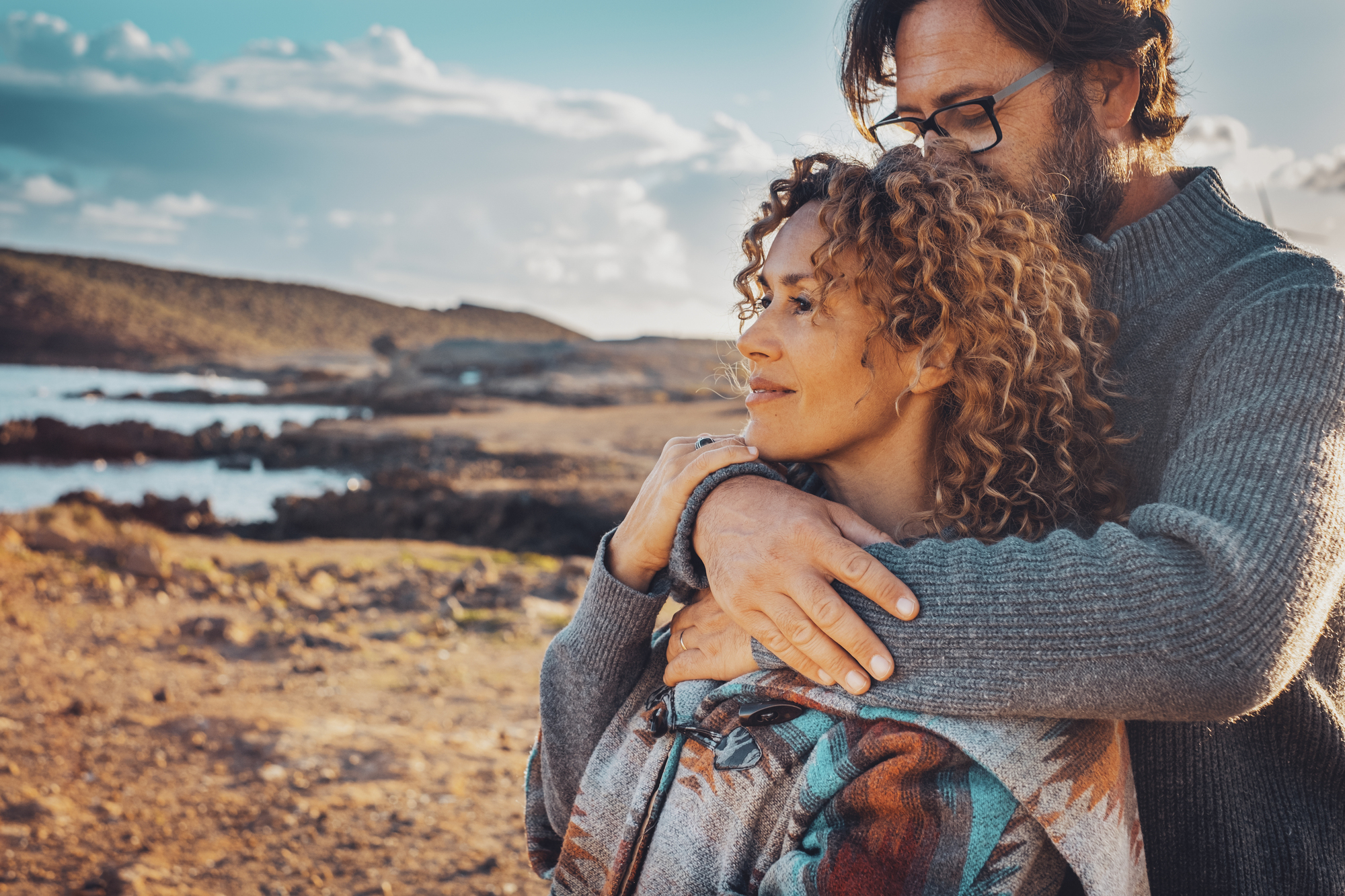 A couple stands close together on a rocky beach with clear blue skies and water in the background. The man, in glasses and a gray sweater, hugs the woman from behind. The woman, with curly hair, smiles contently, both looking towards the horizon.