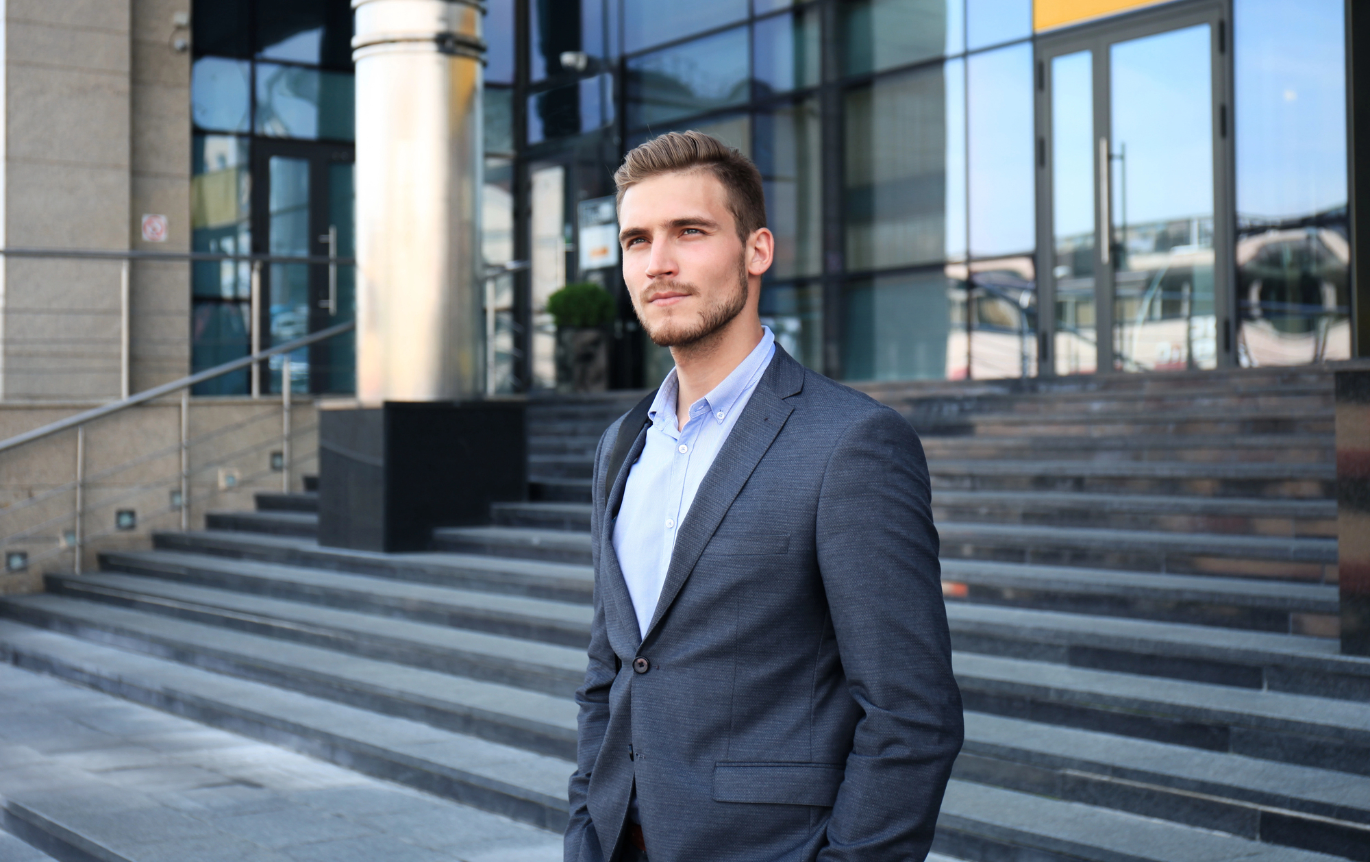 A man in a blue suit stands confidently in front of a modern glass building with a staircase to his right. His hands are in his pockets, and he gazes off into the distance. The background includes metallic and stone architectural elements.