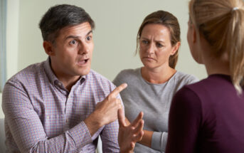 A man with short dark hair gestures emphatically with a finger raised while talking to a younger woman, who is turned away from the camera. Another woman, sitting between them, looks concerned. All are indoors, seated in a casual setting.