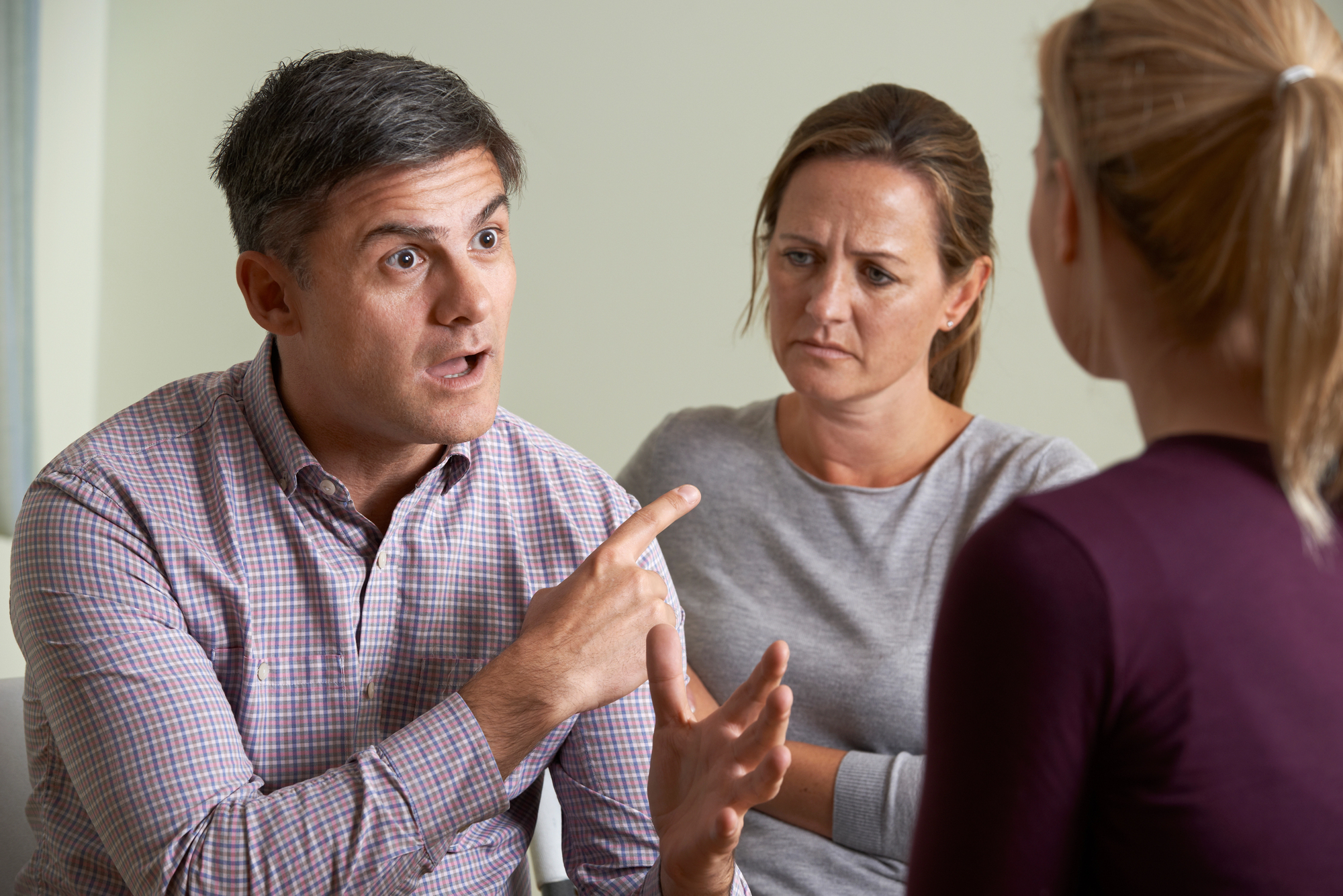 A man with short dark hair gestures emphatically with a finger raised while talking to a younger woman, who is turned away from the camera. Another woman, sitting between them, looks concerned. All are indoors, seated in a casual setting.