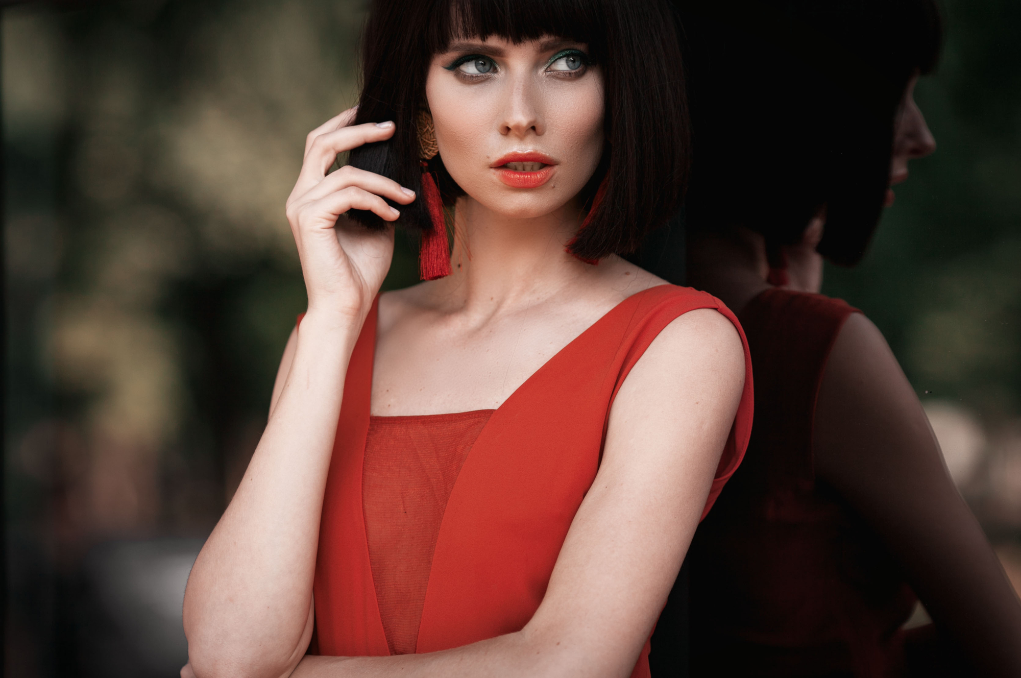 A woman with short dark hair and bold red lipstick is wearing a sleeveless red dress. She poses with her hand near her face, standing against a reflective surface that shows her profile. The background is softly blurred with shades of green.