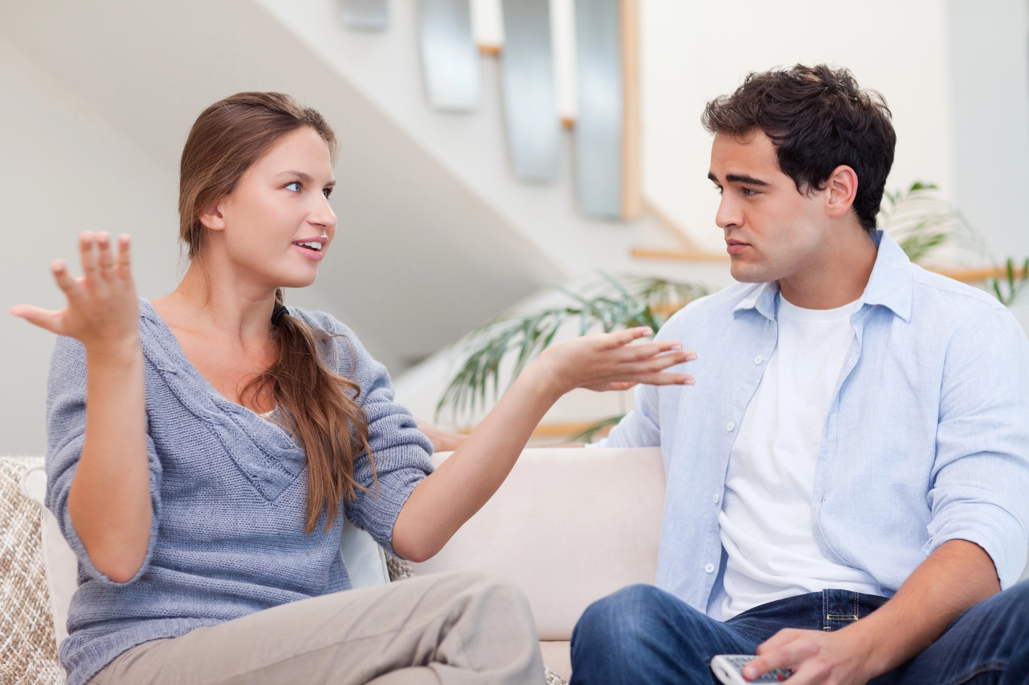 A woman with long brown hair in a gray sweater gesturing with both hands while speaking to a man with short dark hair in a light blue shirt. They are seated on a couch in a modern living room with a staircase in the background.