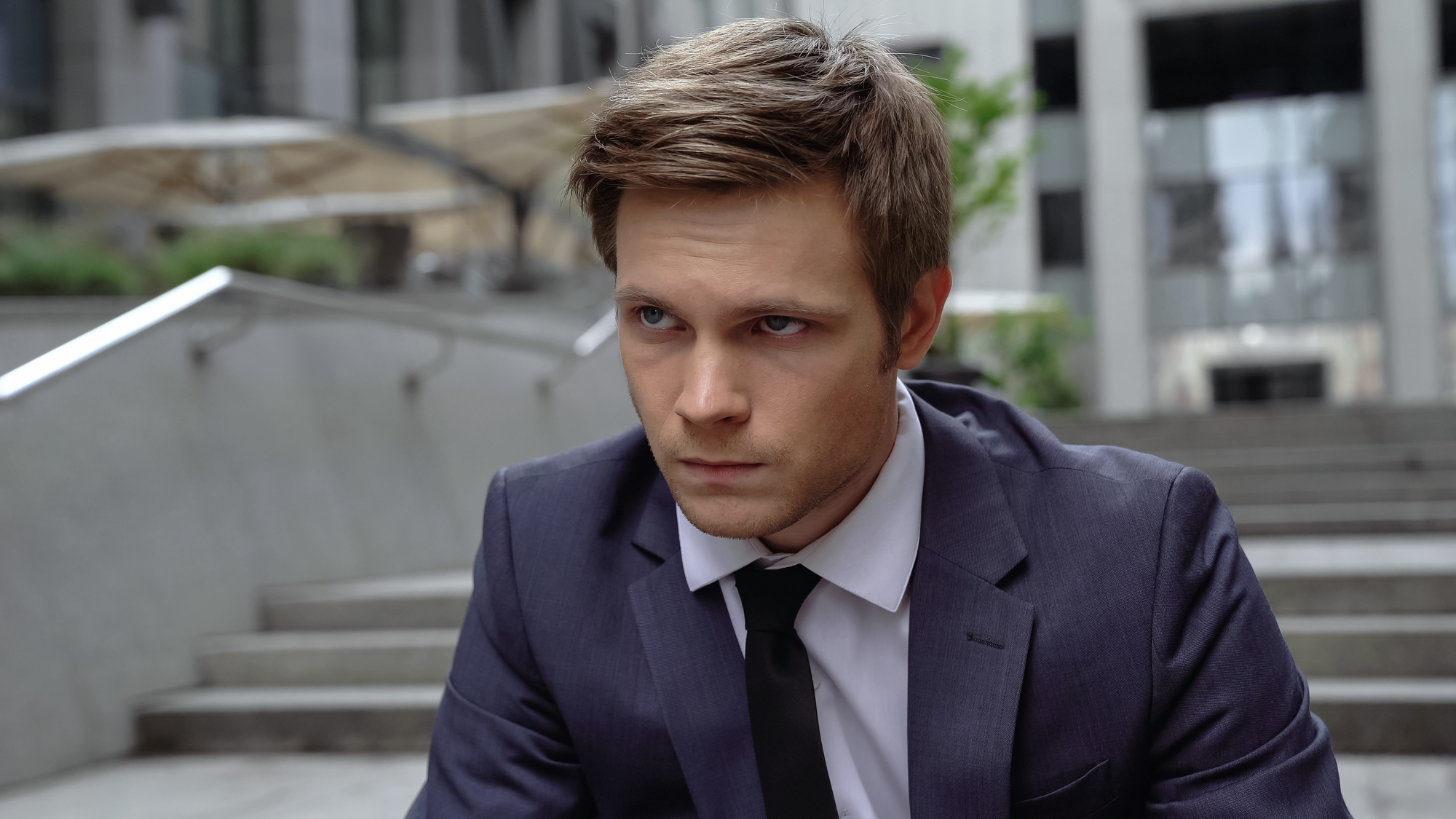 A man with short brown hair in a dark suit and tie sits on a city staircase, looking pensive. The background includes blurred buildings and greenery.