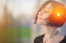 A person with short, dark hair stands with eyes closed, tilting their head slightly upward, basking in the soft sunlight. The warm light creates a serene and contemplative atmosphere. The background is blurred, emphasizing the calm and peaceful expression.