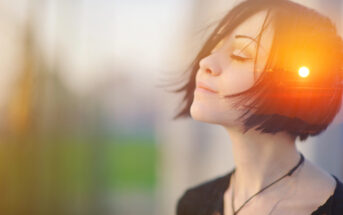 A person with short, dark hair stands with eyes closed, tilting their head slightly upward, basking in the soft sunlight. The warm light creates a serene and contemplative atmosphere. The background is blurred, emphasizing the calm and peaceful expression.