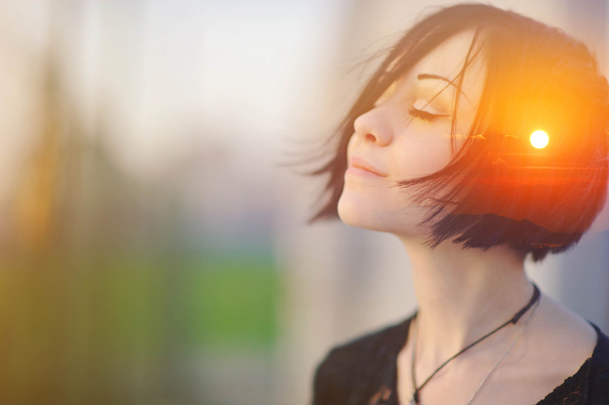 A person with short, dark hair stands with eyes closed, tilting their head slightly upward, basking in the soft sunlight. The warm light creates a serene and contemplative atmosphere. The background is blurred, emphasizing the calm and peaceful expression.